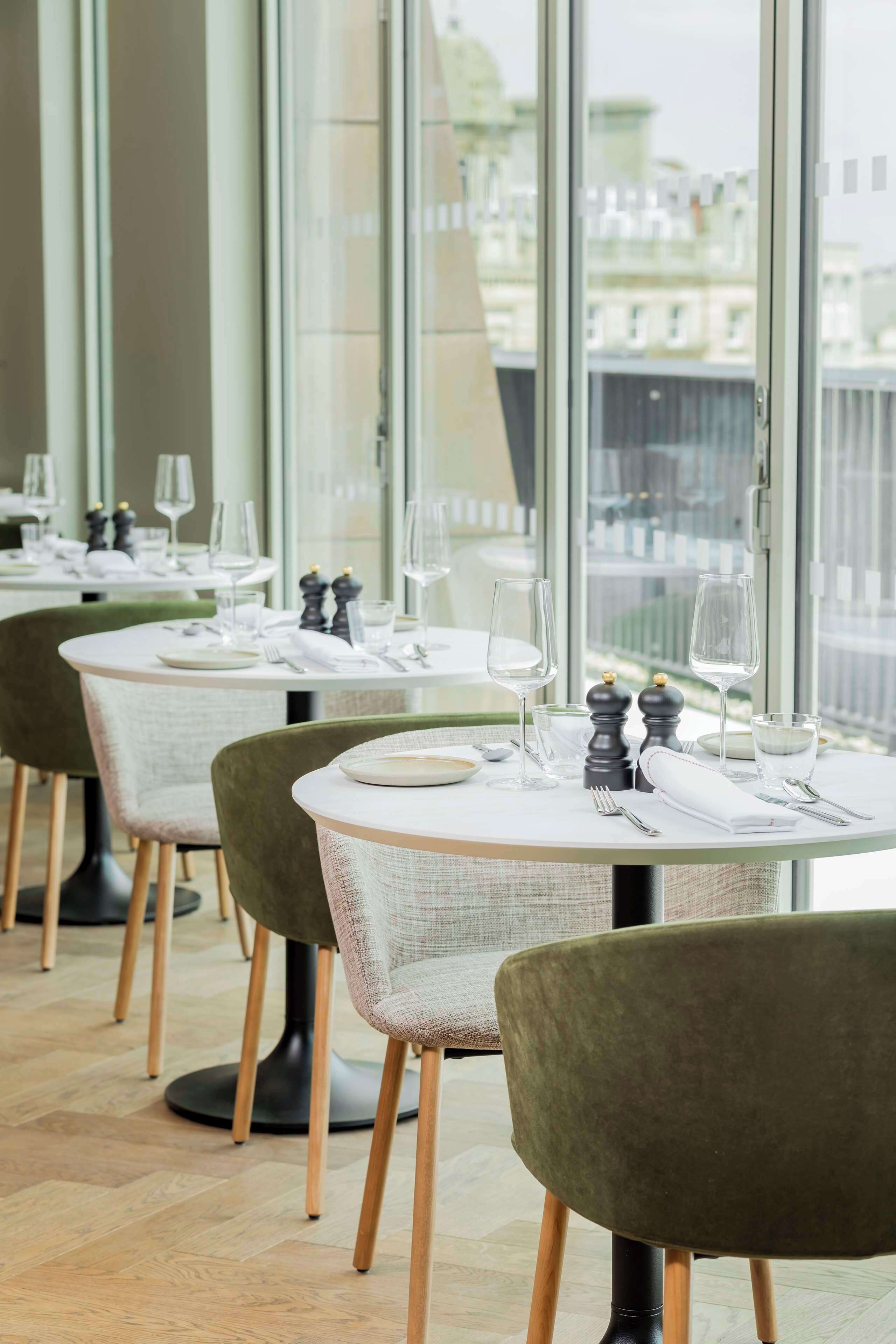 A restaurant with round tables, green chairs, and a view of buildings through large windows.