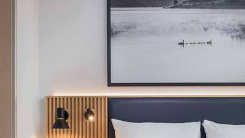 A modern hotel room with a large bed, a bedside table with a lamp, and a wall-mounted artwork of a lake scene.