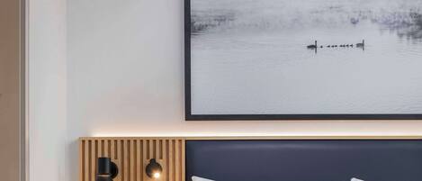 Una habitación de hotel moderna con una cama grande, una mesita de noche con lámpara y una obra de arte colgada en la pared que representa un paisaje lacustre.