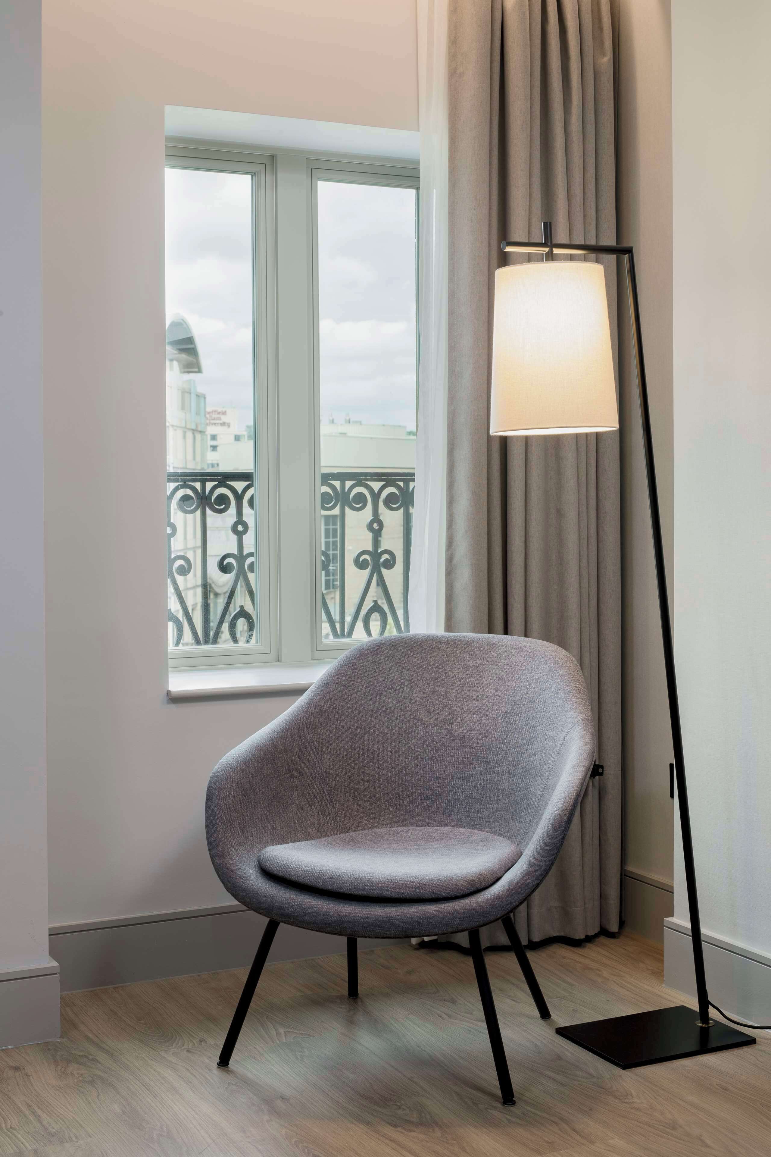 Une chambre moderne avec un fauteuil gris, une lampe sur pied et une fenêtre donnant sur un immeuble et un balcon.