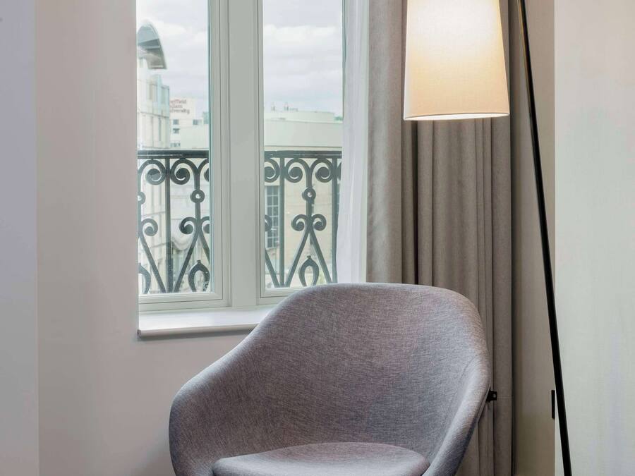 Une chambre moderne avec un fauteuil gris, une lampe sur pied et une fenêtre donnant sur un immeuble et un balcon.
