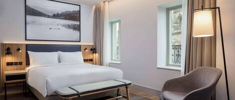 Una habitación de hotel moderna con cama, mesitas de noche, una silla, un ventanal grande y una lámpara de pie.