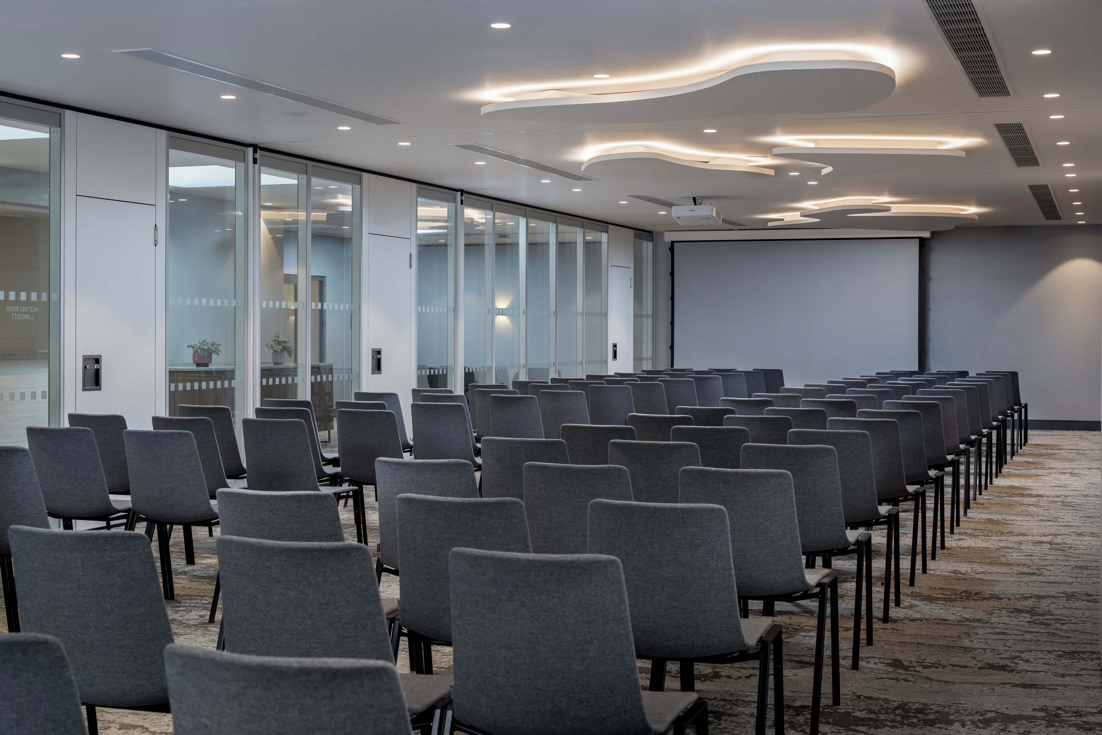 A conference room with rows of grey chairs, a white ceiling with recessed lighting, and a large screen at the front.