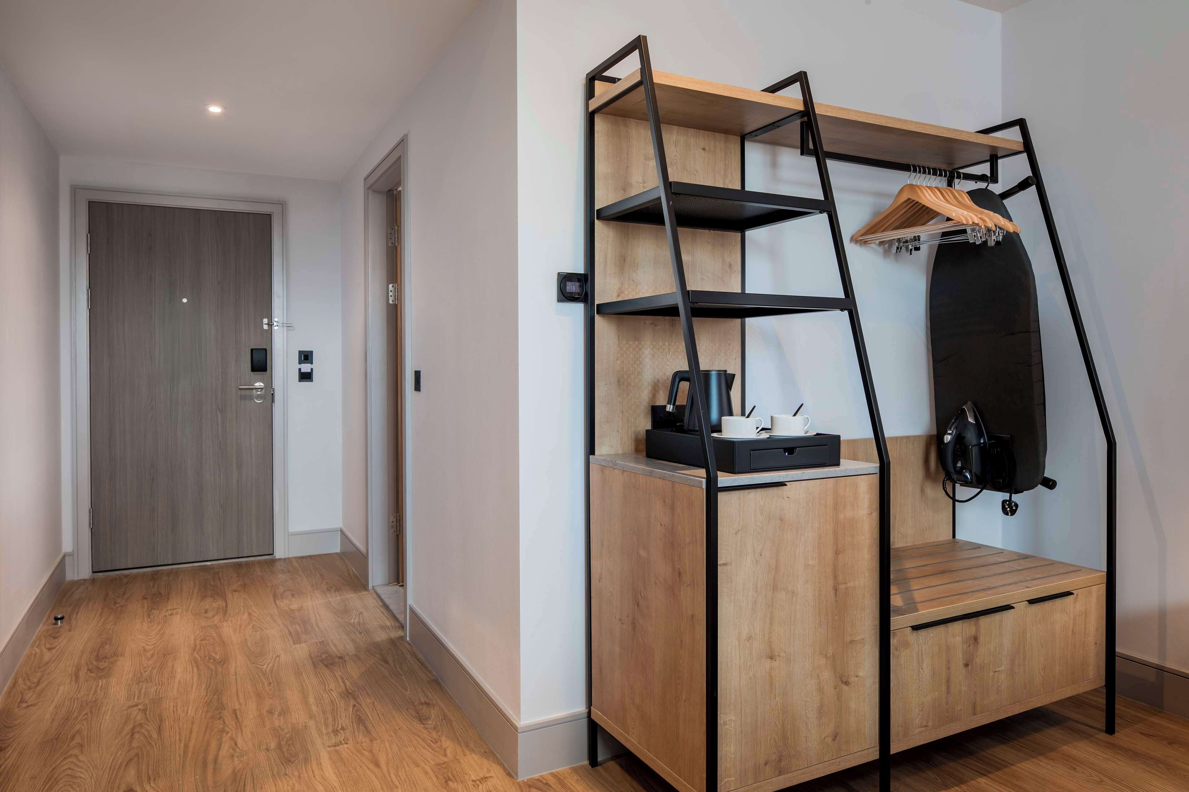 A compact room with a wooden wardrobe, a small desk, and a hanging shelf with a coat rack.