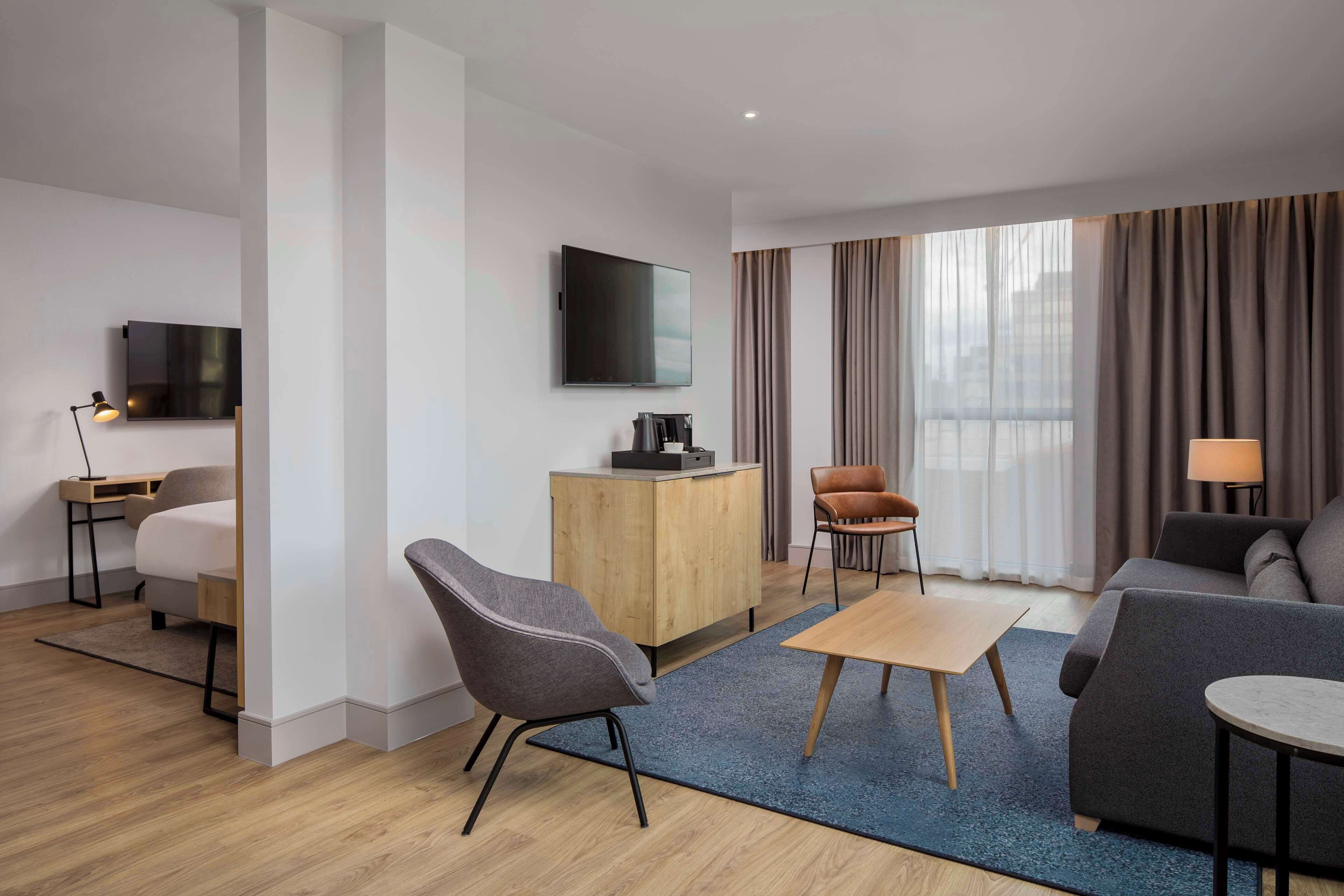 A modern hotel room with a flat-screen TV, a sofa, a coffee table, a desk, and a chair.