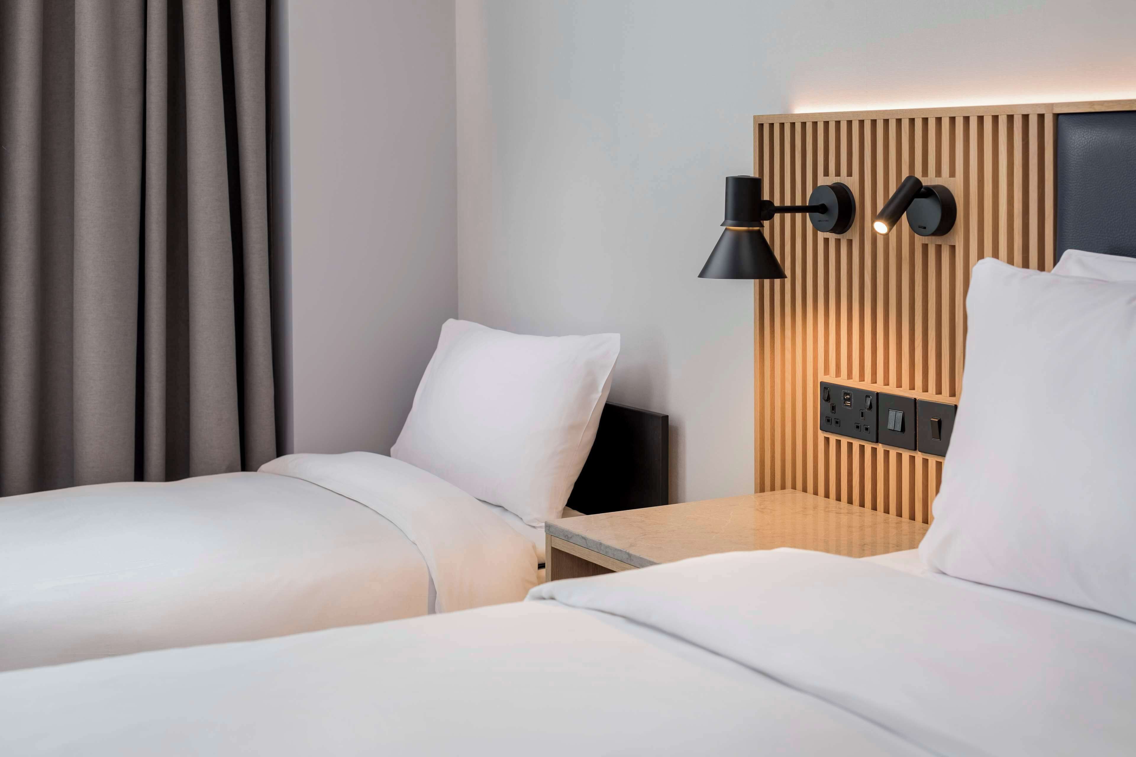 Hotellihuone, jossa on kaksi sänkyä, puinen pääty, yöpöytä ja seinään kiinnitetty lamppu.