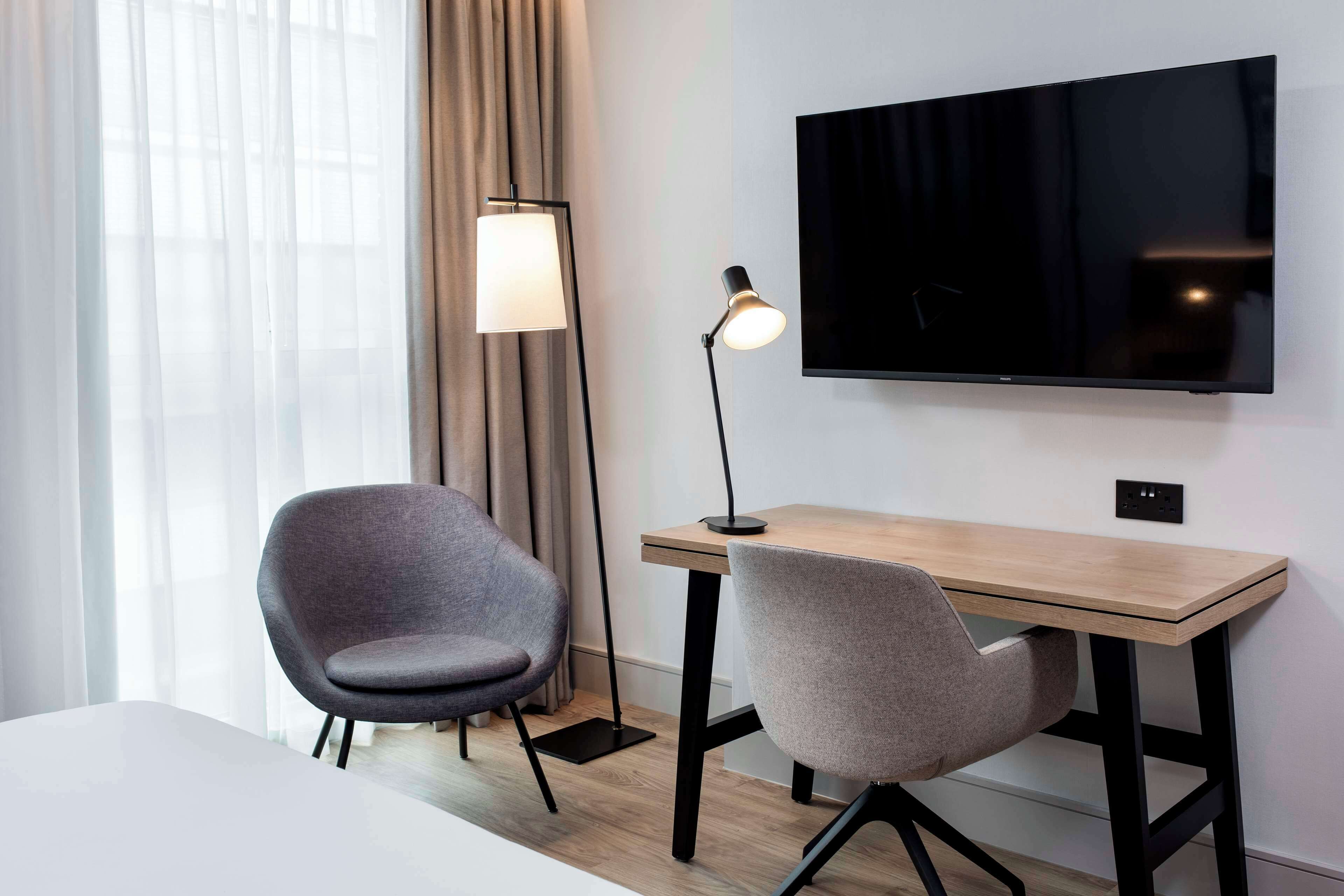 A hotel room with a wooden desk, two grey chairs, a flat-screen TV mounted on the wall, a floor lamp, and a window with sheer curtains.