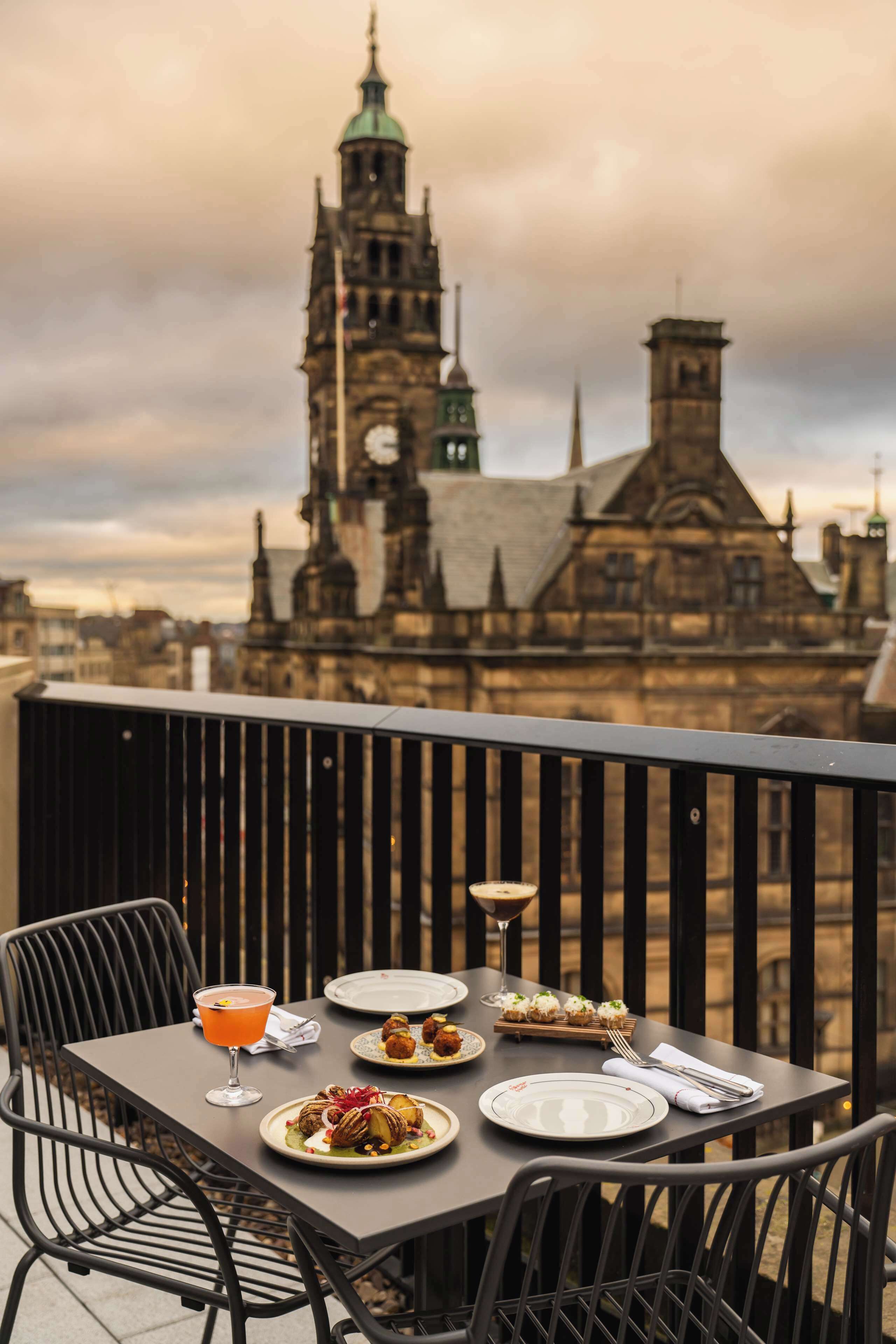 A table set for dinner with plates, cutlery, and a drink on a balcony overlooking historic buildings.