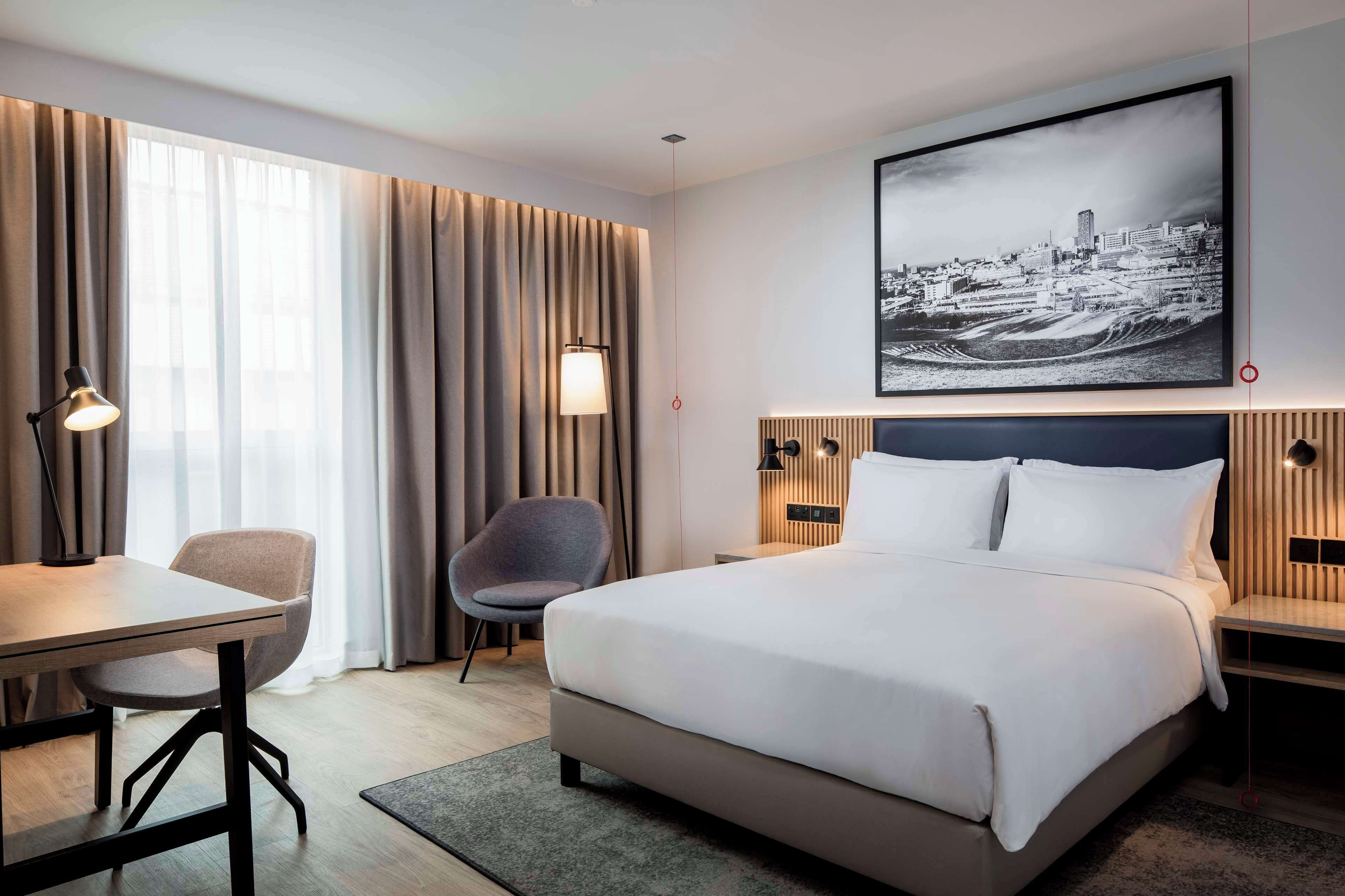 A modern hotel room with a large bed, a desk, a chair, and a cityscape painting on the wall.