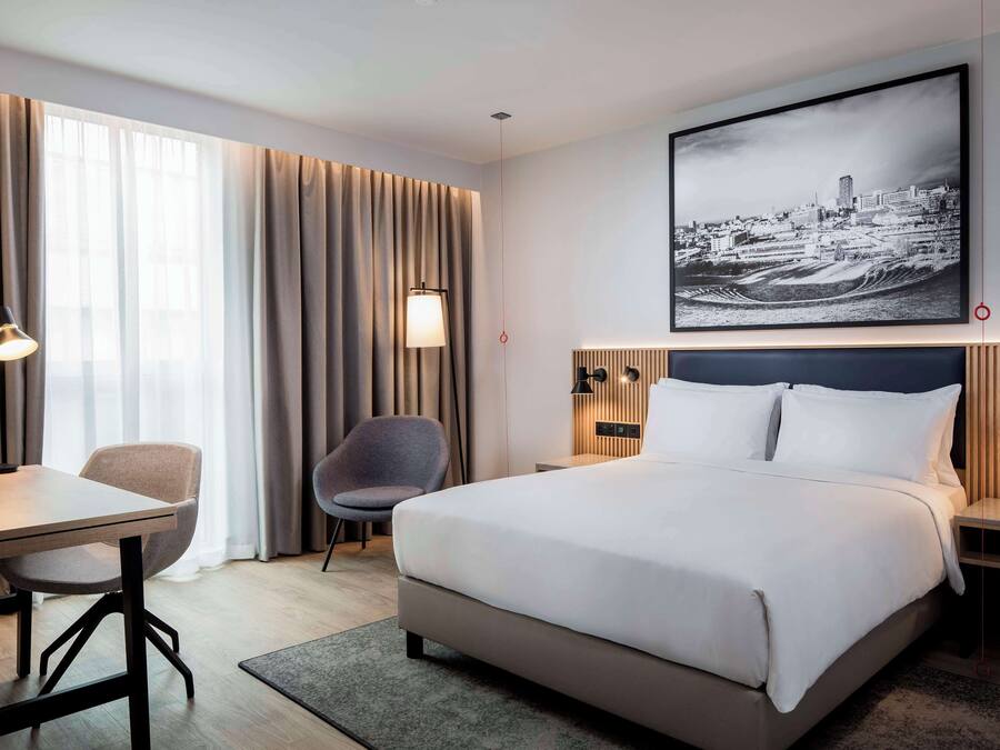 Quarto de hotel moderno com uma cama grande, uma mesa, uma cadeira e uma pintura com vista para a cidade.
