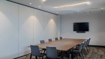 Un salón de conferencias con una larga mesa de madera, sillas grises, iluminación empotrada en el techo y un televisor de pantalla plana montado en la pared.