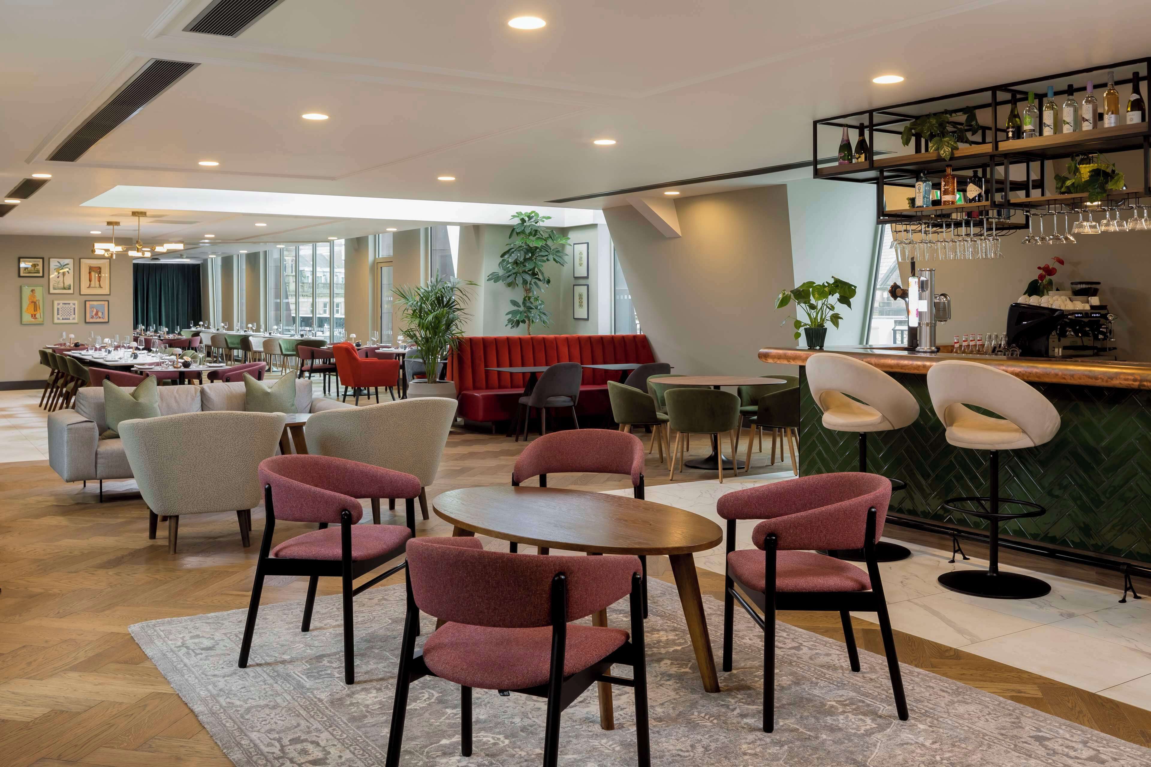Un espace repas moderne avec des tables rondes et des chaises à revêtement rouge, un coin bar avec un mur à motifs chevrons verts, et de grandes fenêtres qui laissent entrer la lumière naturelle.