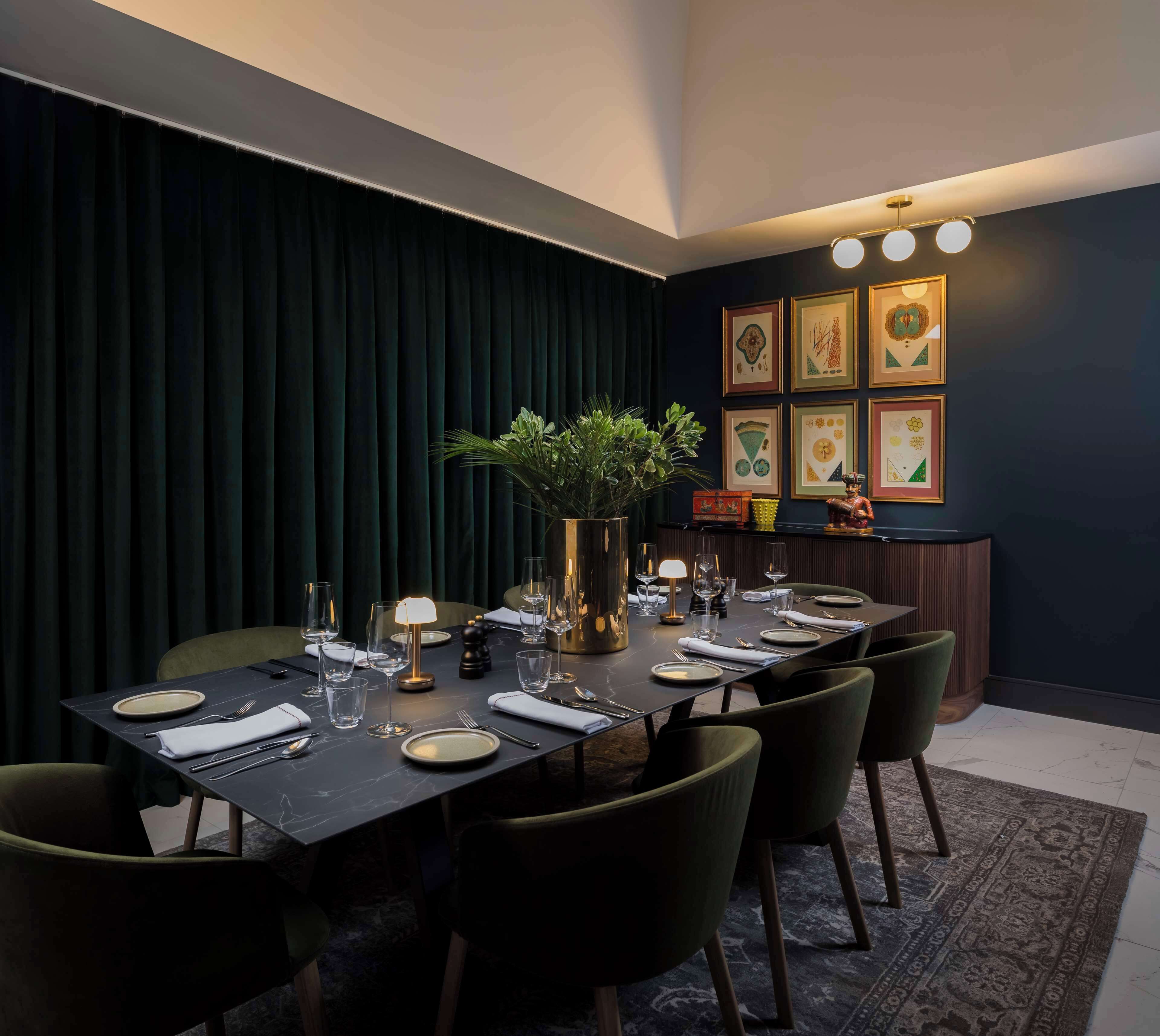 Un coin repas avec une table de couleur sombre, dressée pour six personnes, entourée de chaises. On aperçoit un vase avec une plante sur la table, et la pièce est agrémentée d’un mur foncé orné d’œuvres d’art encadrées.