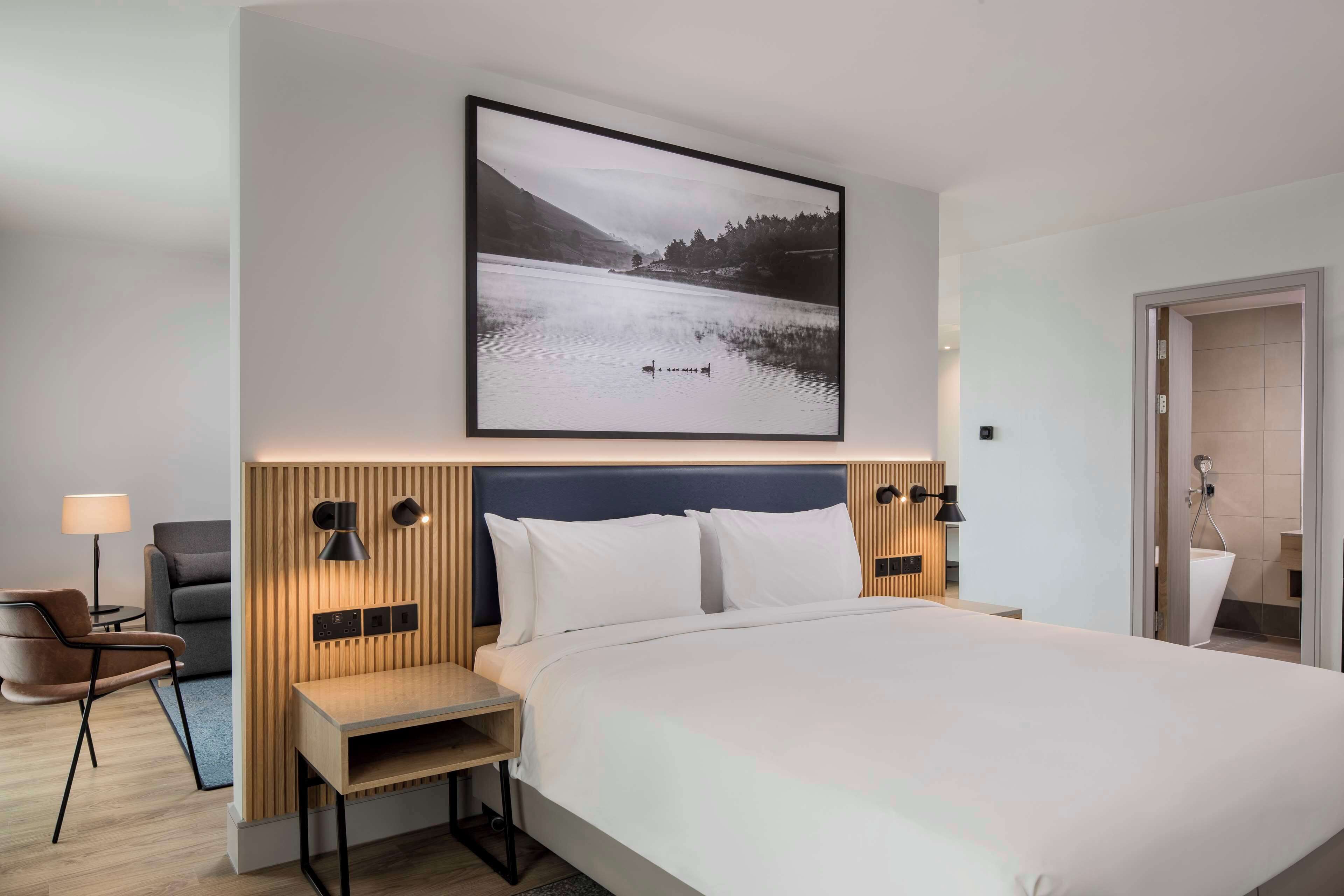 A modern hotel room with a large bed, a wooden headboard, a nightstand, a chair, a desk, and a framed picture of a lake.