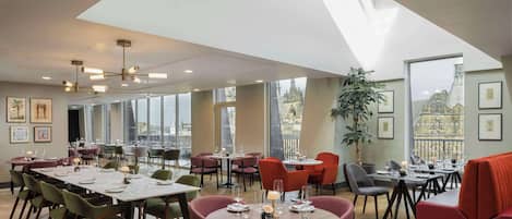 A spacious dining area with multiple tables and chairs, large windows offering city views, and a modern ceiling design.