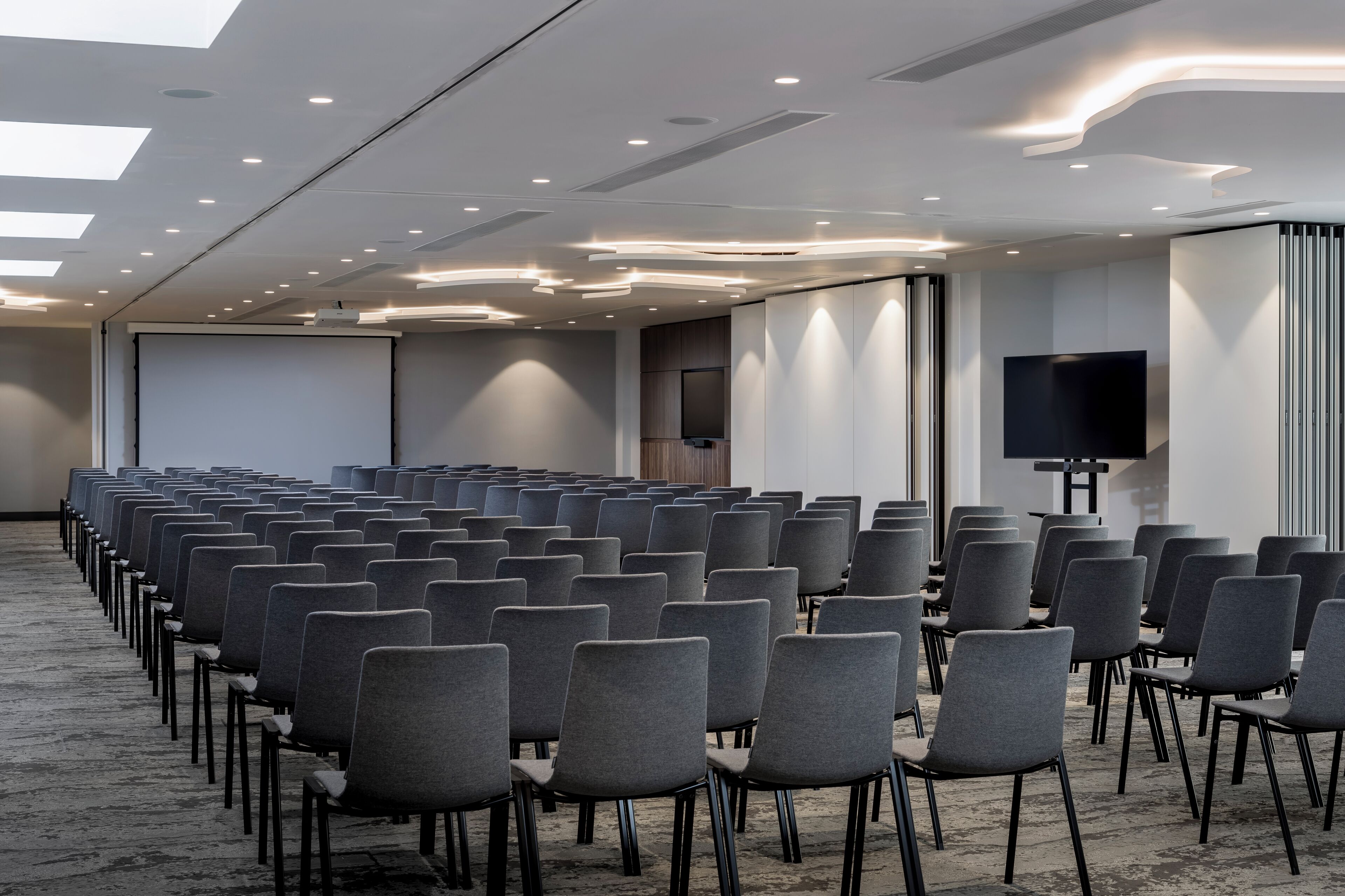 Un salón de conferencias con filas de sillas grises, una pantalla grande y un televisor de pantalla plana sobre un soporte.