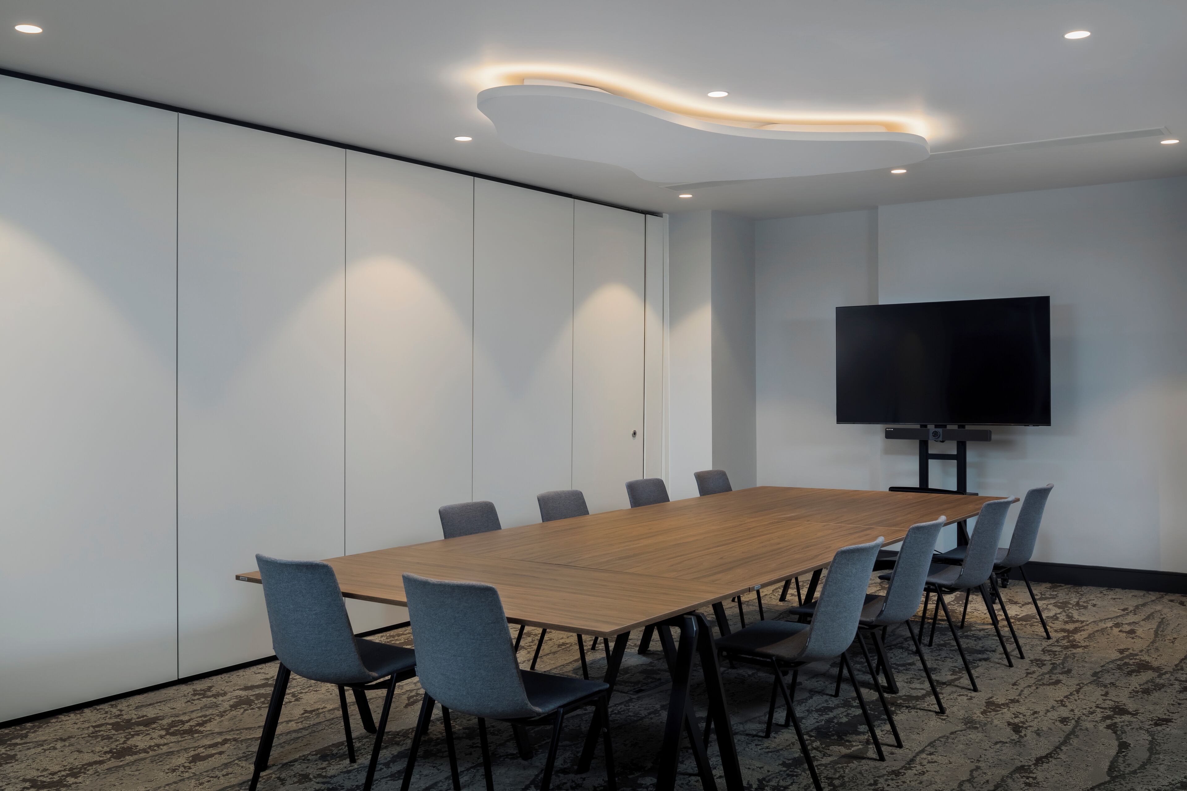 A conference room with a long wooden table, six grey chairs, a flat-screen TV on the wall, and recessed ceiling lighting.