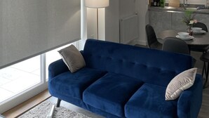 A modern living room with a blue sofa, a dining area with a table and chairs, and a kitchen in the background.