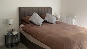 A neatly made bed with a brown bedspread and two pillows, a bedside table with a lamp, and a radiator in the background.