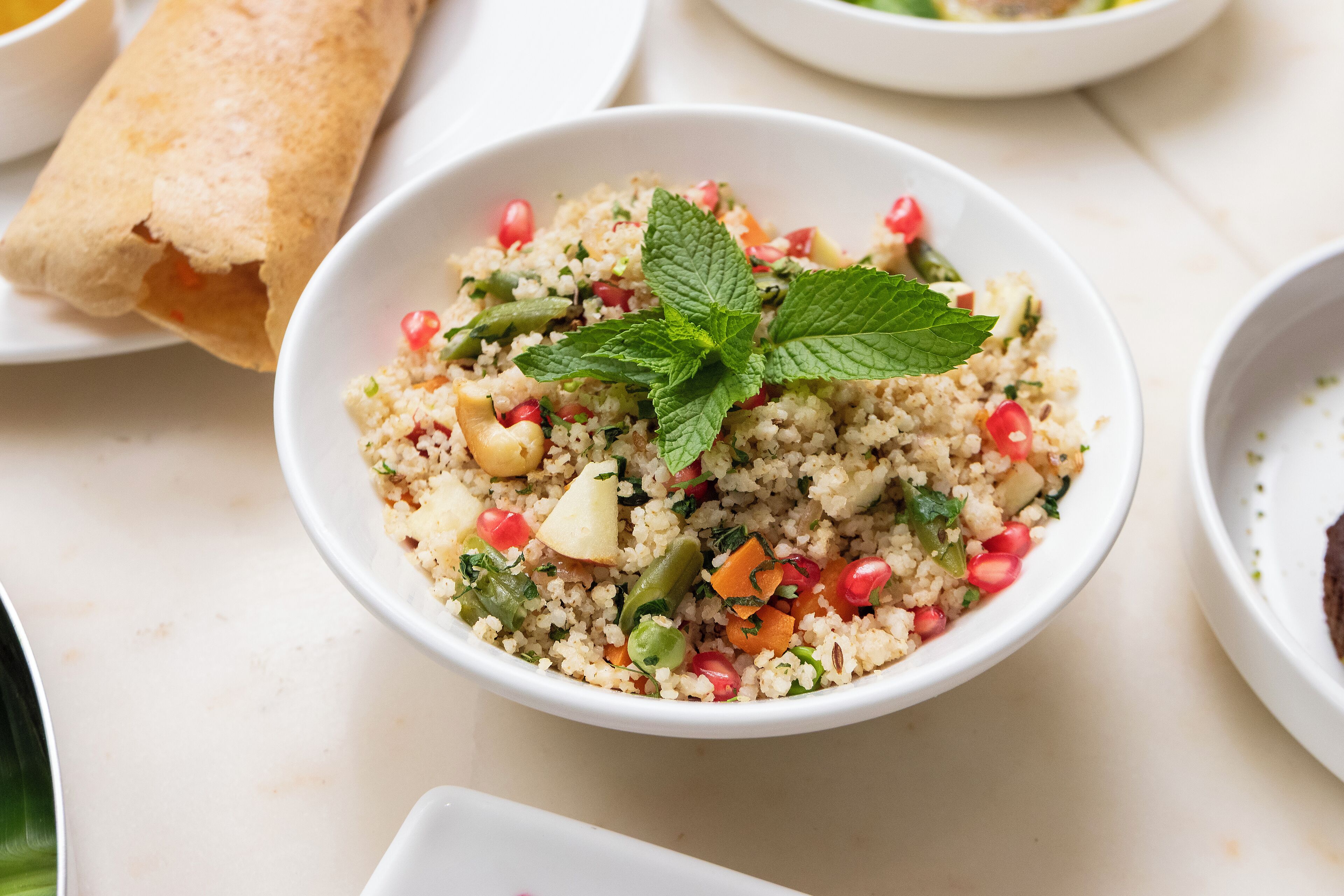 A bowl of couscous salad with vegetables and pomegranate seeds, garnished with mint leaves.
