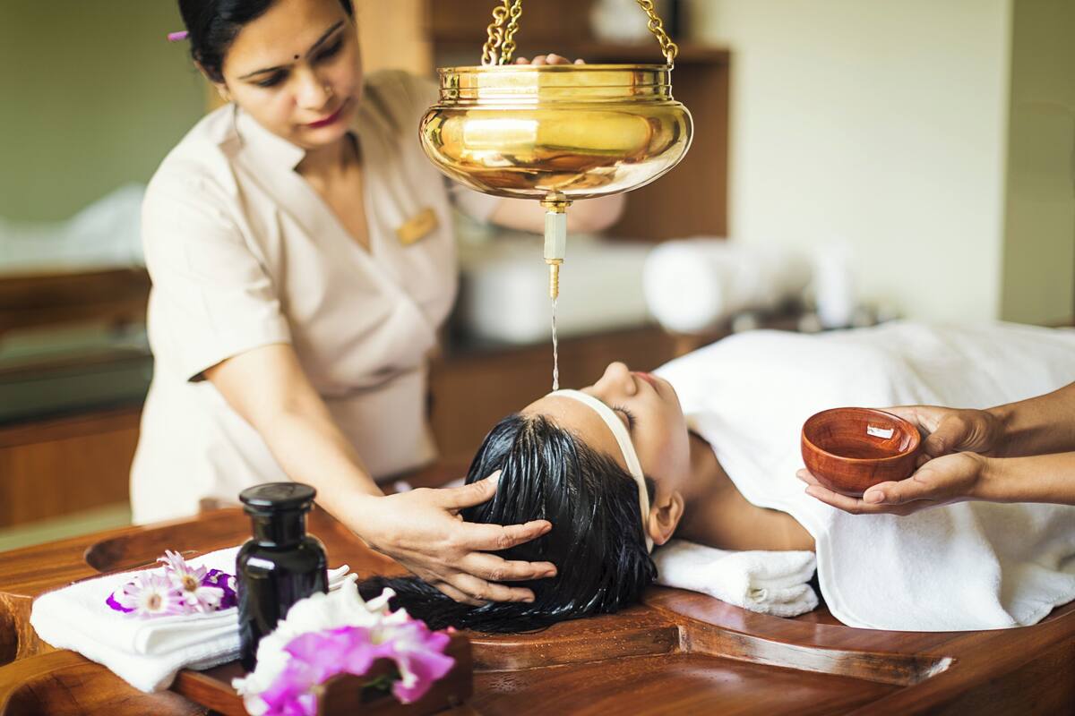 A person receiving a head massage with a golden oil container and a small bowl, in a room with a wooden massage table and a bottle of oil.
