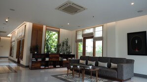 A spacious lobby with a grey sofa, a wooden bookshelf, and a large window with a view of greenery. - Naad Wellness (Sonepat)