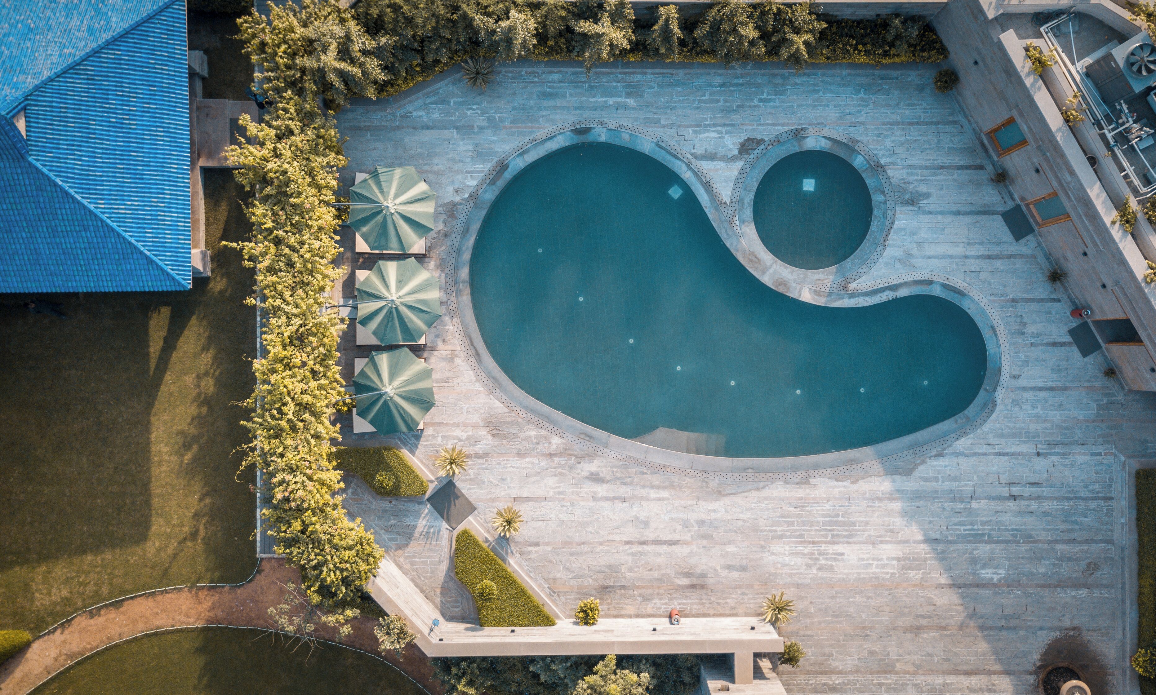 An aerial view of a swimming pool with a unique shape, surrounded by a stone deck and shaded by umbrellas.