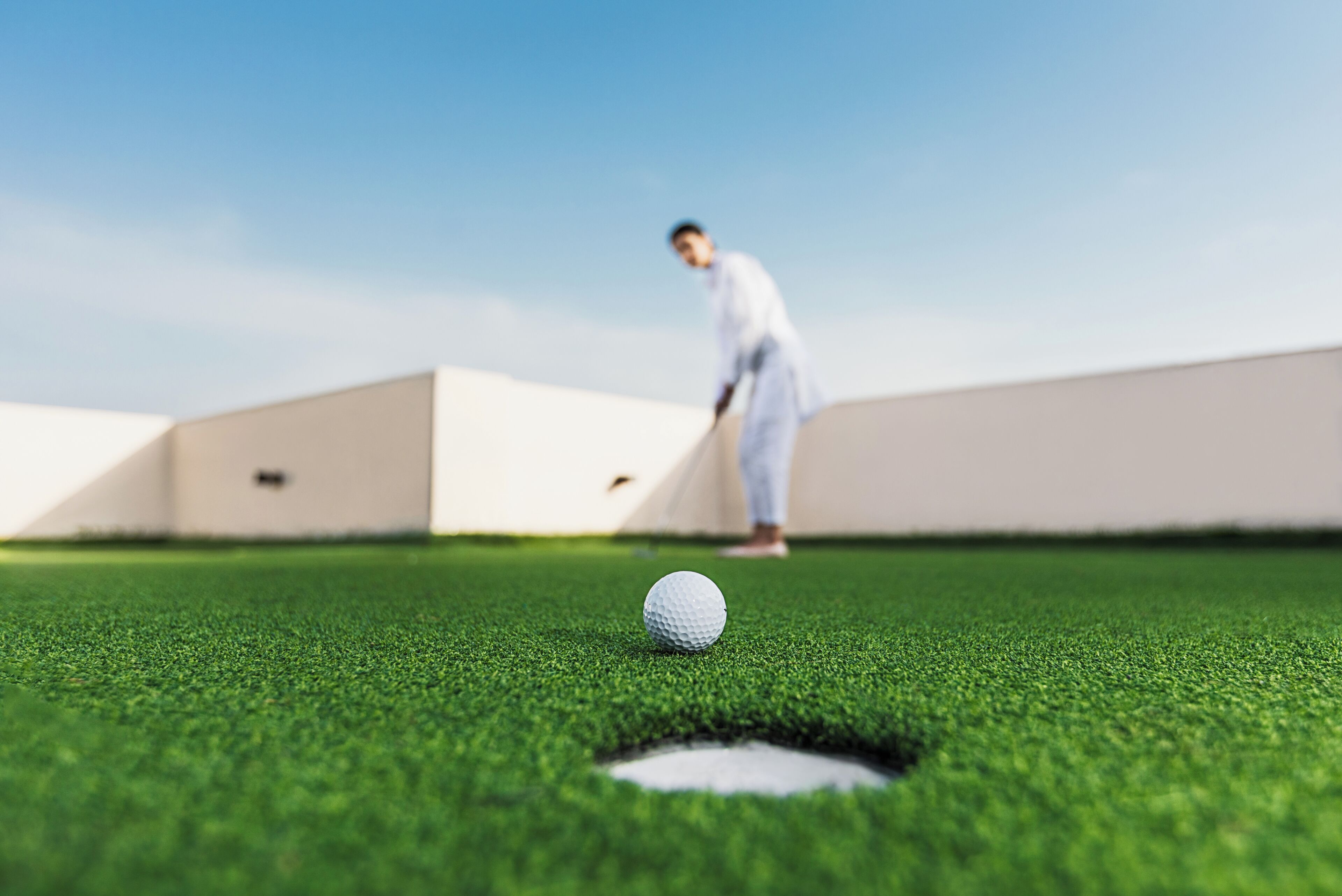 A golf ball on a green with a hole, a person in the background, and a clear sky.