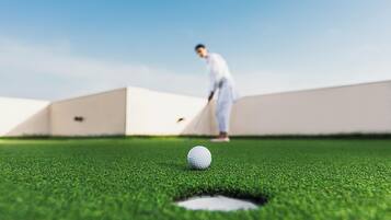 A golf ball on a green with a hole, a person in the background, and a clear sky.
