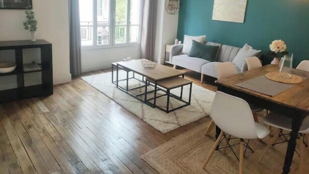 A modern living room with a sofa, coffee table, dining table, and a large window. - Charmant Appartement au Pied D'un Parc Arboré (Clichy)
