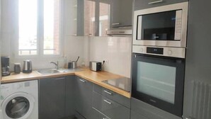 A compact kitchen with grey cabinets, a built-in oven, a microwave, a washing machine, and a window providing natural light. - Charmant Appartement au Pied D'un Parc Arboré (Clichy)