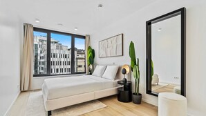 A modern bedroom with a large bed, a window with a city view, a large mirror, and two potted plants.