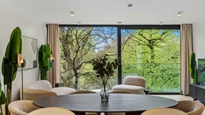 A modern dining area with a round table, beige chairs, and a large window with a view of trees.