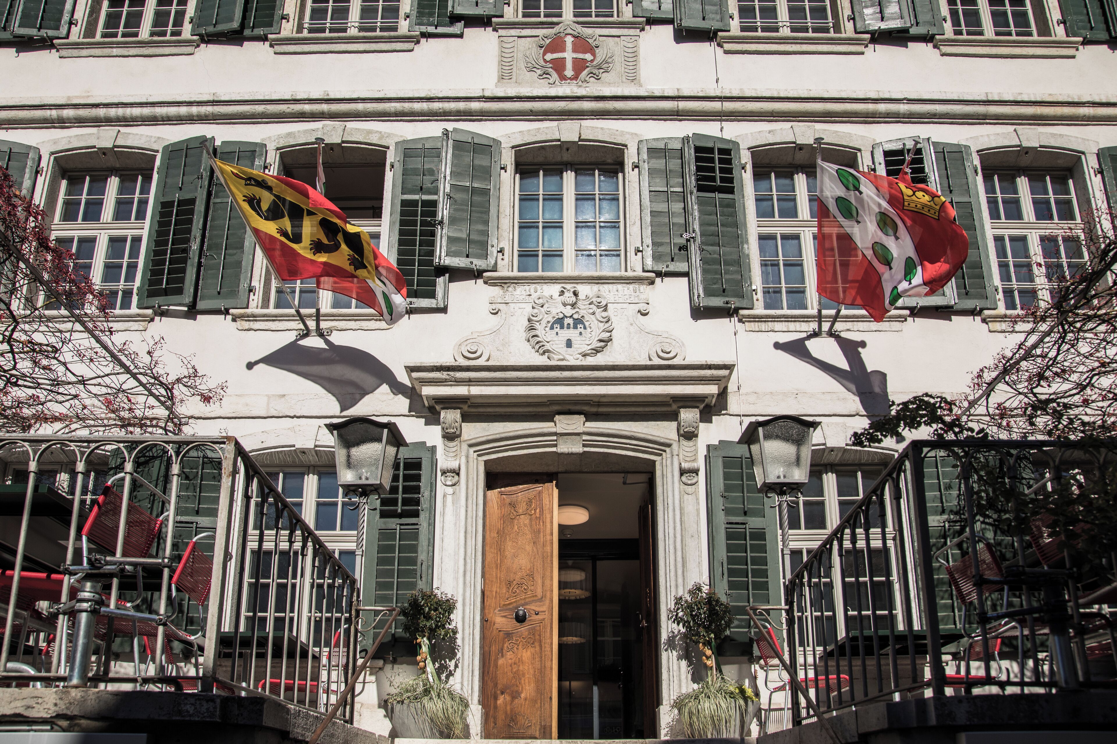 A building with a central entrance, multiple windows with green shutters, and flags on either side.