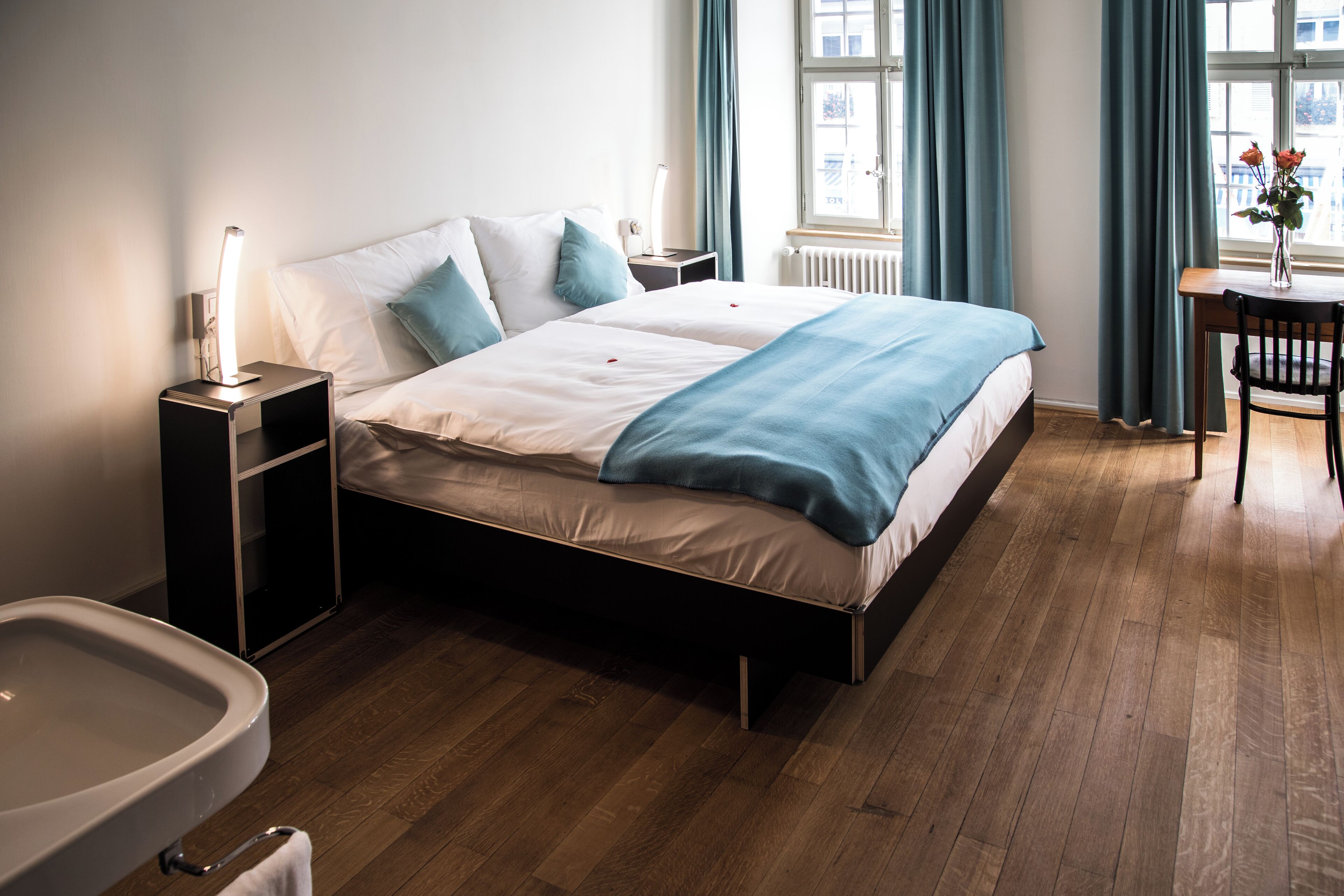 Une chambre d’hôtel moderne avec un plancher en bois, un lit avec une literie blanche et bleue, une table de chevet avec une lampe, une petite table avec un vase de fleurs et une vue sur la ville à travers de grandes fenêtres.