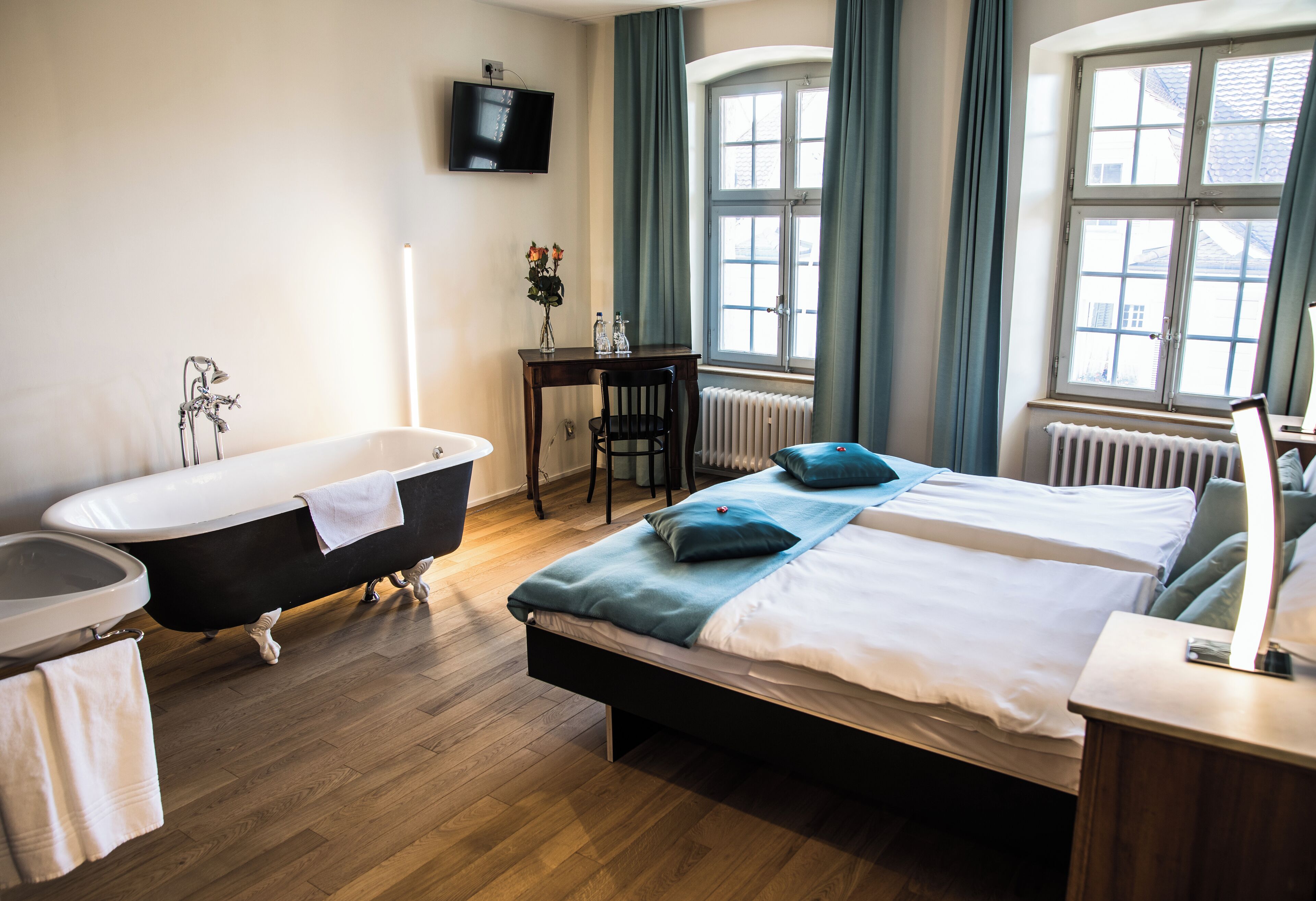 A hotel room with a bathtub, a bed with pillows, a desk, and a window with curtains.