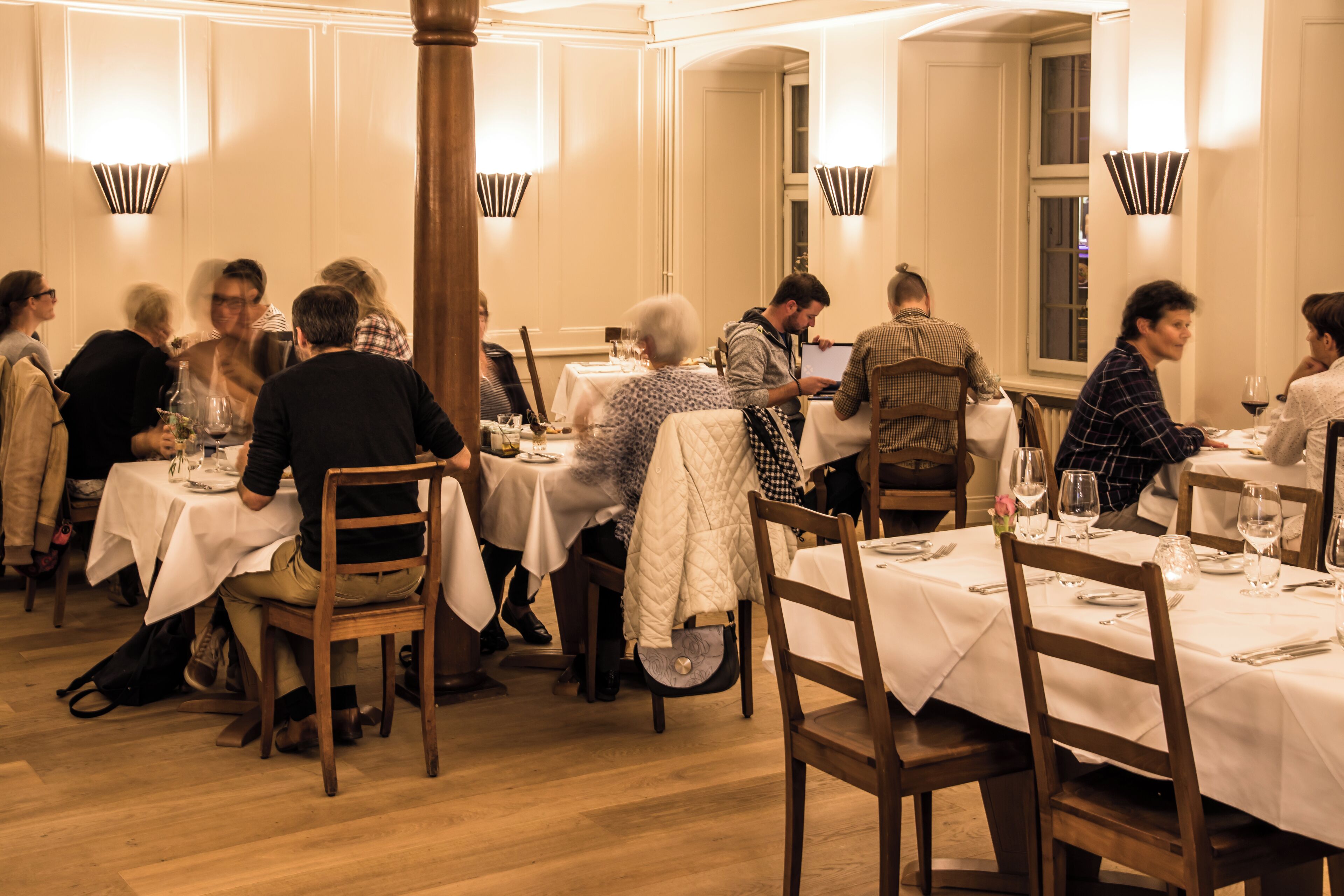 Un restaurante con varias mesas y sillas, pisos de madera y luminarias fijadas a la pared.