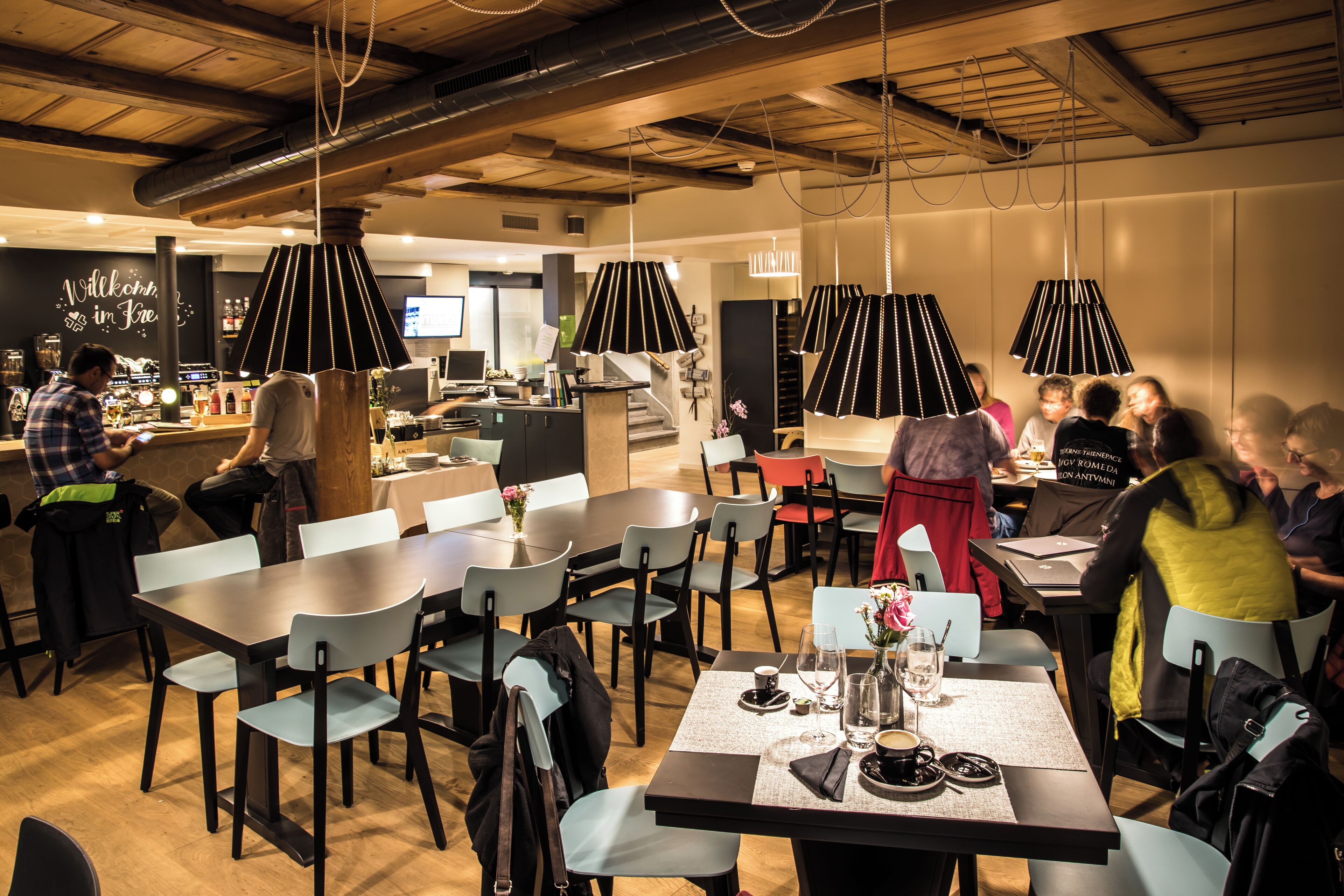 Moderni ravintola, jossa on useita ruokapöytiä ja -tuoleja, baarialue sekä katosta roikkuvia riippuvalaisimia.