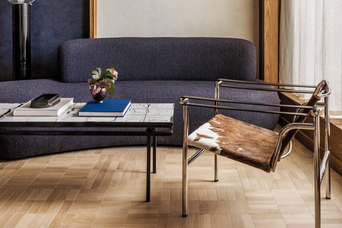 A modern living room with a dark blue sofa, a metal-framed chair, a coffee table with books and a vase, and a large mirror on the wall.