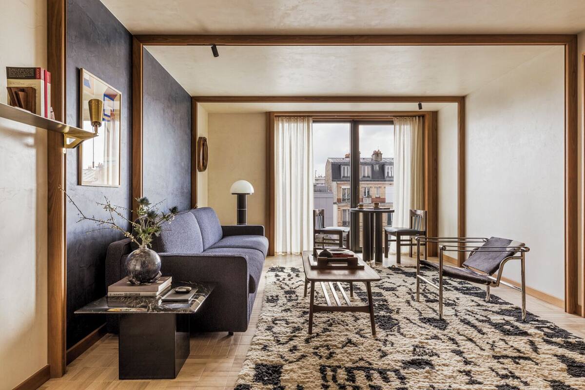 A modern living room with a sofa, coffee table, and armchairs, a large window with curtains, and a view of buildings outside.