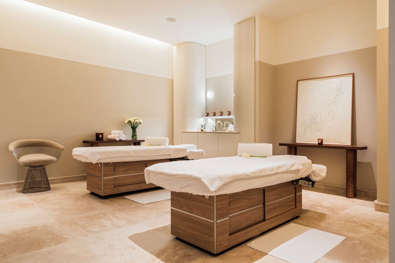 Ein Spa-Raum mit zwei Massagetischen, einem Sessel, einem Spiegel und einem gerahmten Kunstwerk an der Wand.