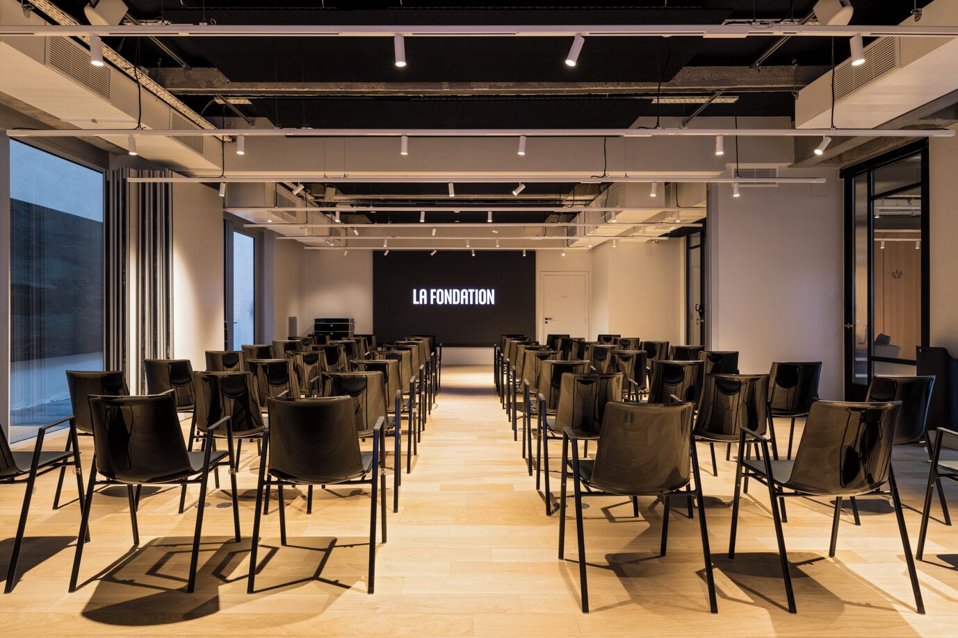 A conference room with rows of chairs facing a screen displaying "LA FONDATION".