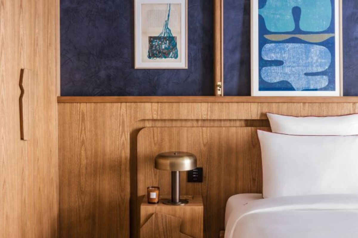 A neatly made bed with white linens and a wooden headboard. There is a small bedside table with a lamp and a candle. The wall is adorned with abstract art.
