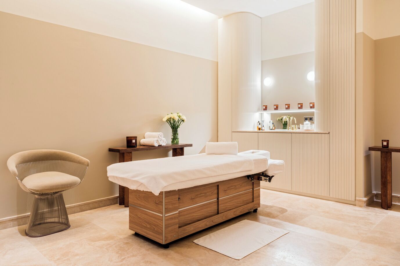 A modern spa room with a wooden massage table, a cushioned chair, a small table with a vase of flowers, and a mirror with a lighted vanity.