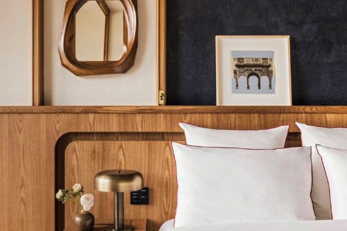 A hotel room with a wooden headboard, white pillows, a bedside table with a lamp, a framed picture of an archway, and a mirror.