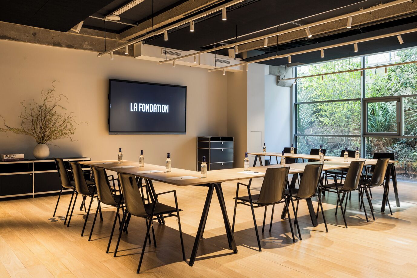 Ett modernt konferensrum med en stor skärm som visar texten "LA FONDATION", bord i trä, svarta stolar och ett stort fönster med utsikt över grönområden.