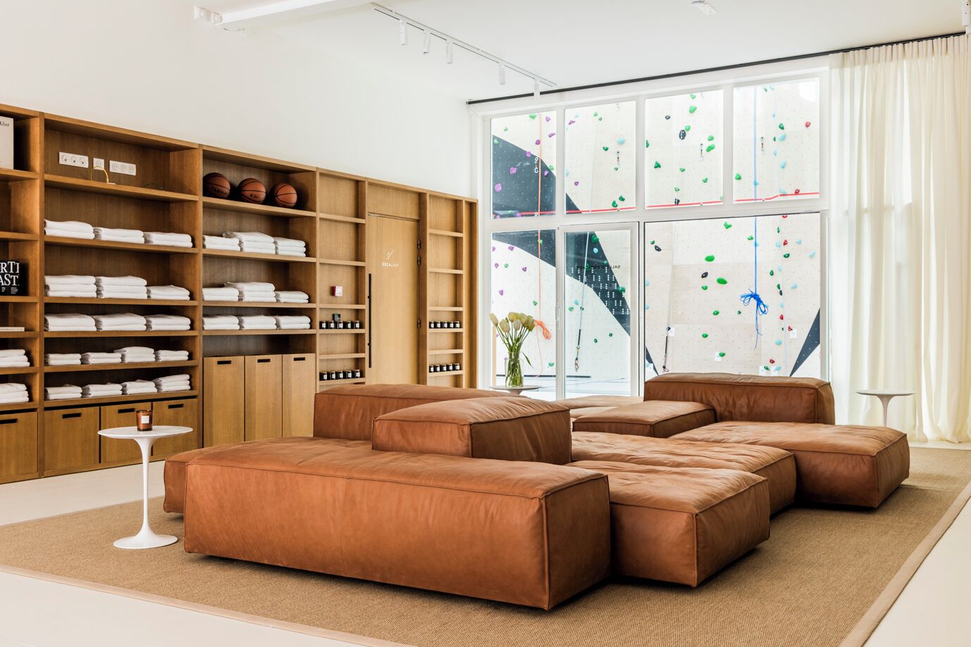A modern living room with a sectional sofa, wooden shelving with towels, and a large window with a climbing wall view.
