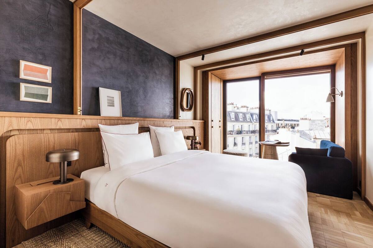 A hotel room with a large bed, wooden headboard, a nightstand, a desk, and a view of the city through large windows.