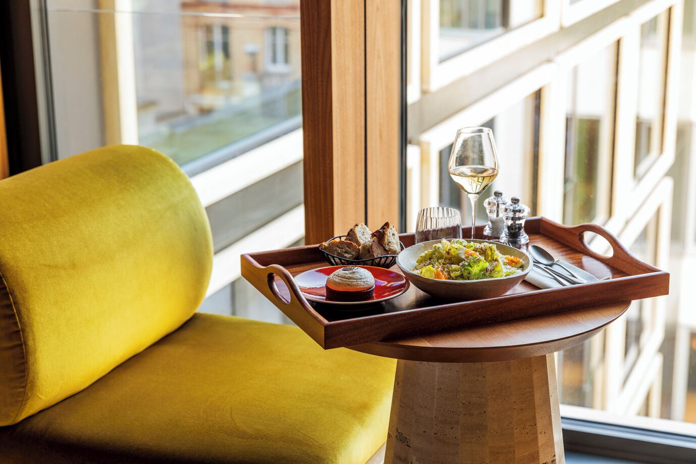 Una silla amarilla junto a una ventana con una bandeja que contiene una ensalada, pan y una copa de vino.