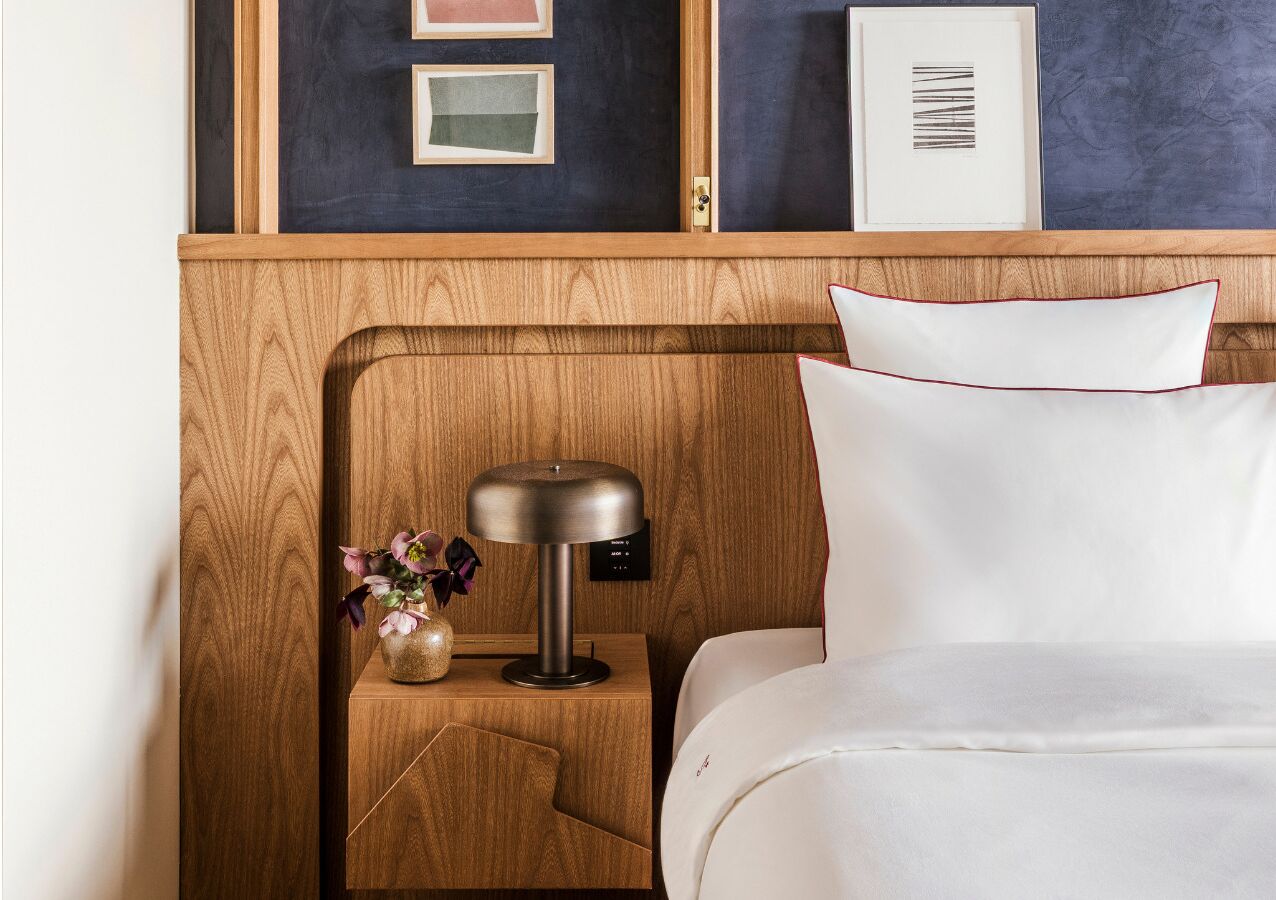 Una cama bien hecha con ropa blanca, una mesita de noche de madera con una lámpara moderna, y un jarrón con flores.